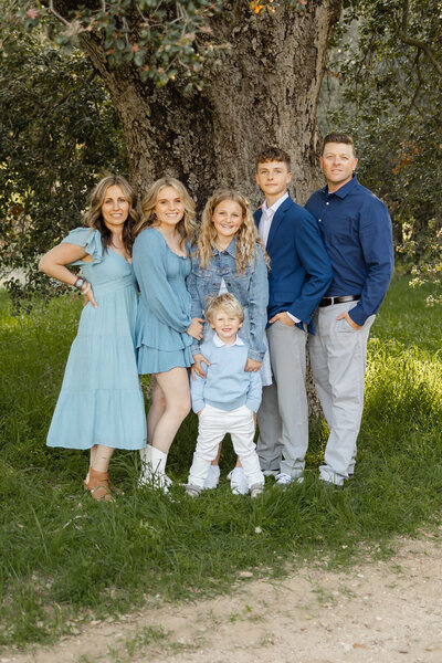 Family standing outdoors under a large tree, dressed in coordinated blue and white outfits. Photographed by Yucaipa family photographer Kaitlyn Dawn Photography.