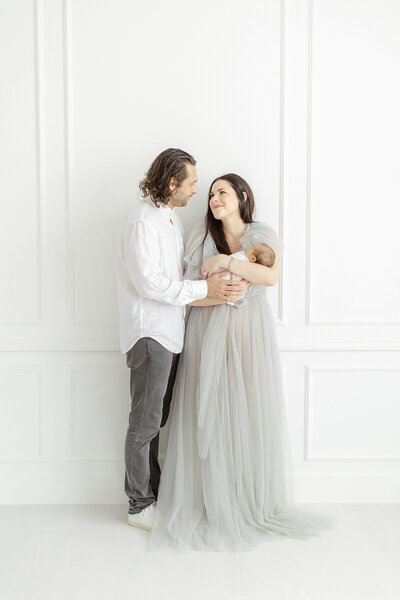 Photo of new parents to a newborn baby boy as they hold each other while they stand in a Dallas/Fort Worth photography studio posing for their babies newborn session.