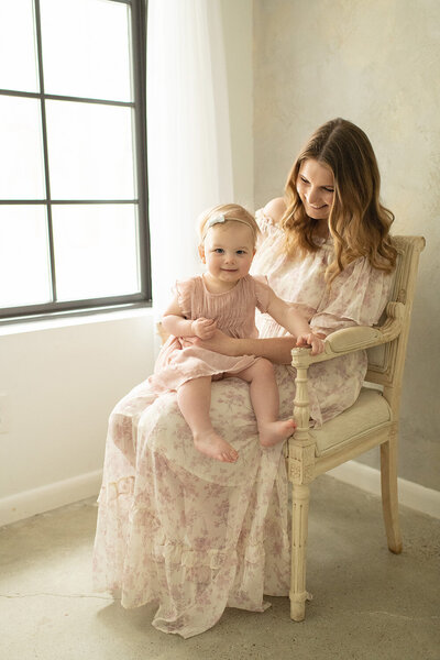 Mother in a floral dress sits on a vintage chair, smiling at her toddler on her lap. A sweet motherhood session by Moments in Light Photography.