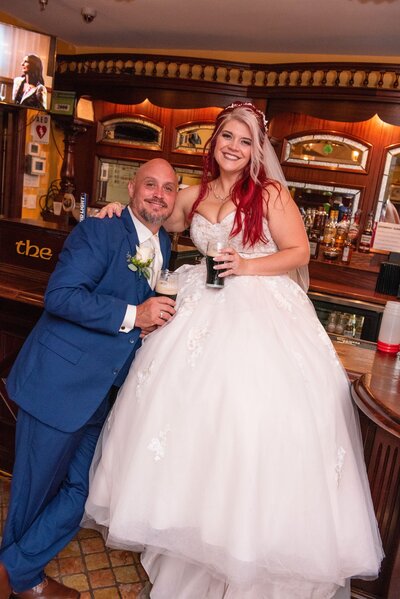 Bald man smiling with a beer in his hand stands next to bride with red and blonde hair in a fluffy princess gown and a beer in her hand.
