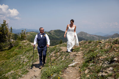 Bride and groom hike along a scenic mountain ridge in Montana during their adventure elopement. The bride wears a white wedding dress and hiking boots, while the groom, dressed in a navy vest and tie, uses trekking poles on the rocky trail. Rolling mountain peaks stretch into the distance under a clear blue sky.