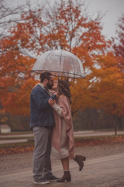 Engagement Couple Photo, the couple are standing under an umbrella, in a gloomy Fall afternoon, with a Fall colored trees in the backdrop