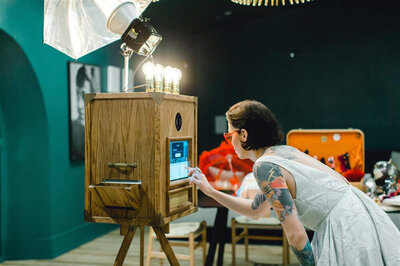 Tattooed guest using vintage wooden photo booth with umbrella lighting during an event.