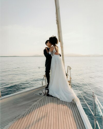 Bride and groom embracing on a sailboat at sunset, with the bride wearing a flowing lace wedding gown