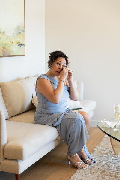 New England Wedding Photographer Pricing | Kelsey Sheehan Photography Timeless Rhode Island Weddings | A woman in a light blue dress sits on a beige sofa, applying makeup. The room is softly lit, conveying a calm and poised atmosphere.