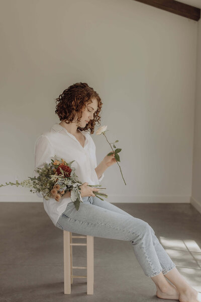 Woman sitting on a stool, holding a bouquet of flowers and smelling a rose