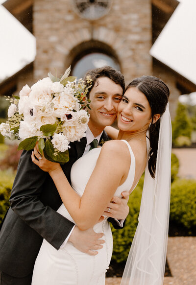 An image showing a Bride and Groom lovingly embracing in front of a chapel and the Bride is holding her bouquet.