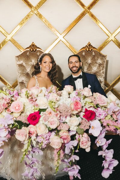 Bride and groom smile behind a pink and purple floral installation at their wedding reception.
