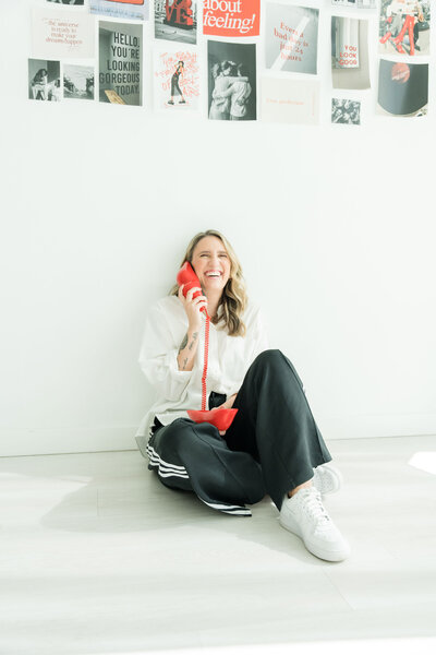 Latina woman business owner sitting on the floor against a white wall, dressed in white blouse and pants, confidently using a red telephone, representing empowerment and website design services for Latina owned businesses.