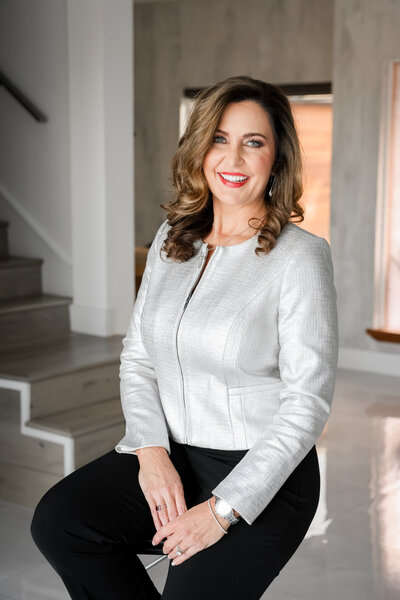 A woman with shoulder-length, wavy hair wearing a silver jacket and black pants sits inside a modern room with a staircase in the background.