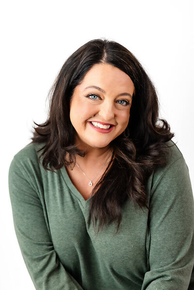 Woman with black hair and a green sweater leans over and smiles playfully into camera.