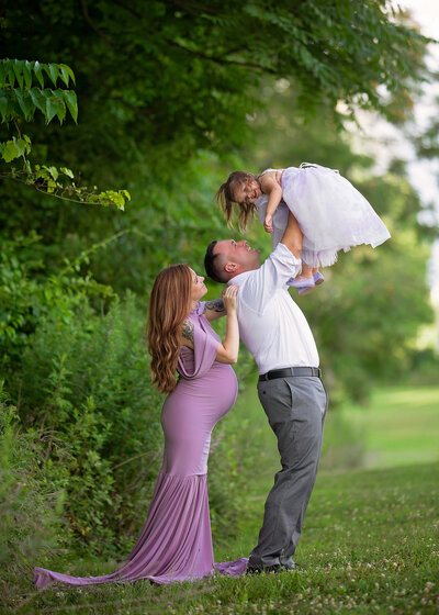 A pregnant woman wearing a lilac gown is standing with her husband who is playfully tossing their daughter in the air
