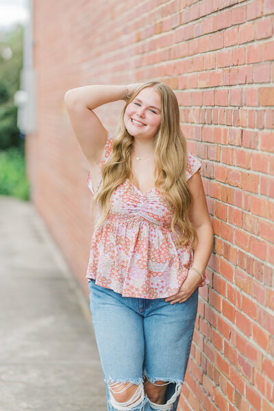 High school senior girl smiling against a brick wall in downtown Raleigh during her portrait session with Lindsey Lambert Photography.