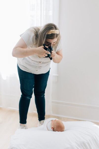 Woman stands taking pictures of newborn baby wrapped in swaddle blanket.