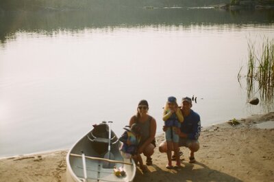 A family crouching in the sand beside a canoe in front of a lake