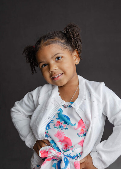 Black girl with curly pigtails in a white dinosaur dress posing with hands on her hips for a preschool portrait with Las Vegas Photographer, Jessica Bowles