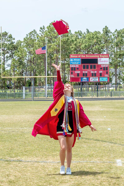 A high school senior in a red gown tossing her graduation cap in the air on the Ashley High School football field in Wilmington, North Carolina. This energetic photo captures the excitement and celebration of graduation, perfect for capturing vibrant senior moments.