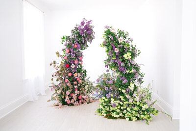 A standalone floral installation indoors, with two curved columns of blooming lilacs, hydrangeas, roses, and greenery arranged symmetrically in a bright white room.