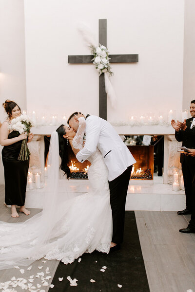 A bride and groom share their first kiss in front of a candlelit fireplace and large cross at SoHo63, a Chandler wedding and event venue. The bride’s long veil cascades to the floor as the groom dips her, surrounded by clapping guests and soft romantic lighting.