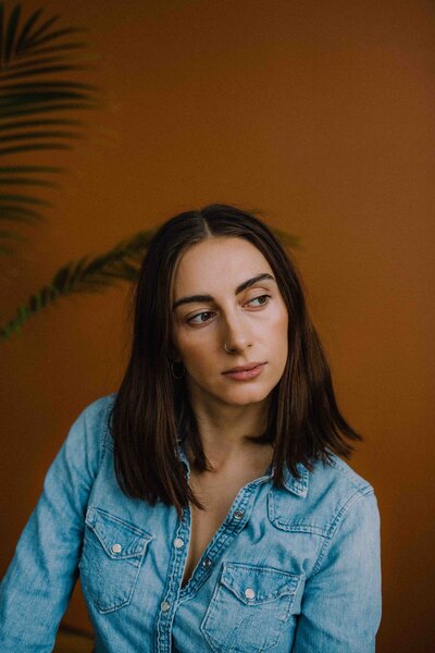 A woman with straight dark hair and a nose ring looks off to the side with a contemplative expression, wearing a light denim shirt in front of a warm terracotta-colored background with a soft palm leaf shadow.