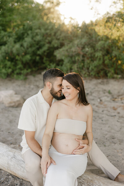 A mom to be in a two piece outfit sitting on a beach with her husband at sunset while smiling down to her bump