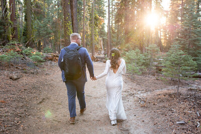 A newlywed couple walks hand in hand down a sunlit forest trail during their Yosemite elopement, with the groom carrying a backpack for their adventure. Captured by Yosemite elopement photographers, this moment embodies the freedom and intimacy of an outdoor wedding.