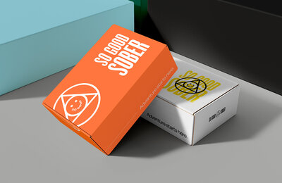 Two branded boxes on a flat surface. The orange box reads “SO GOOD SOBER” in bold white text with a circular smiley logo and “Adventure starts here.” on the side. The white box features the same design in yellow and black. Surrounding boxes in teal, green, and black add a modern, vibrant feel.