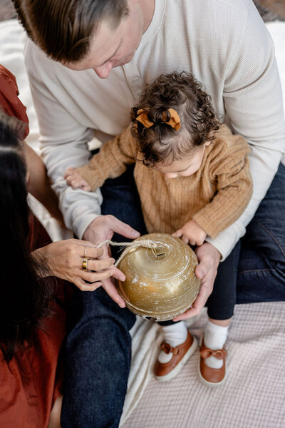 mother and father hold toddler as they show her a large gold Christmas bell ornament while they sit on a white blanket
