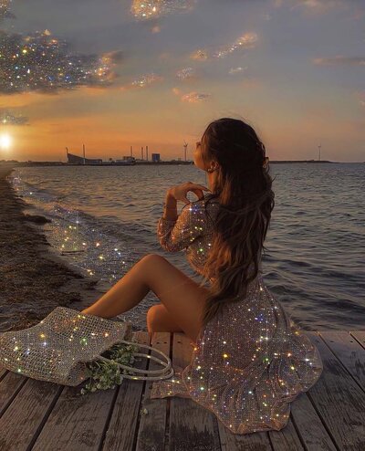 A woman in a sparkling dress sits on a wooden dock by the water, gazing at the sunset. Her bag and the surrounding area shimmer, creating a magical effect. This image is for Debra LeClair's Embodied Style services.