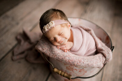 Newborn with headband in a bucket with pink blankets smiling