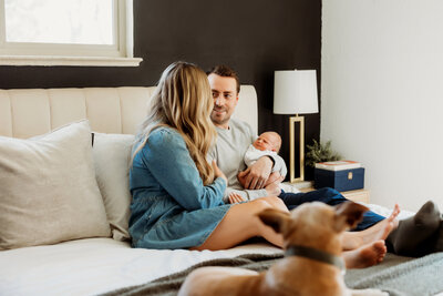 mom and dad sitting on their bed with their puppy and new baby for their at home newborn photo session