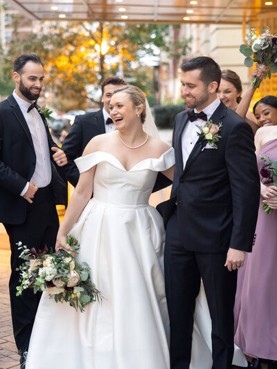 A joyful bride and groom stand together, surrounded by bridesmaids and groomsmen. The bride wears an elegant white gown, holding a bouquet, while the groom is in a tuxedo. The group is outdoors in a festive atmosphere.