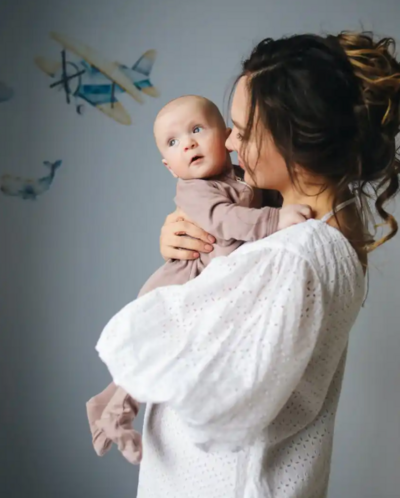 Mother in white dress holding baby in nursery.