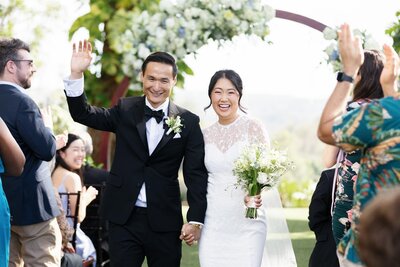 A newlywed couple holding hands and smiling as they walk back up the aisle 