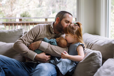 mom standing by window holding toddler girl and kissing her cheek as girl closes her eyes and leans in towards mom
