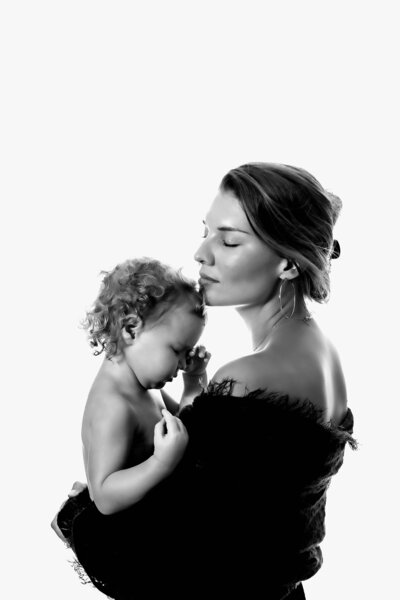 A very modern, artistic black and white motherhood photo captured by Mobile, AL Family photographer. The mom is resting her head on the top of her toddler's head as the toddler rubs her eyes sleepily. The mom closes her eyes as she takes in the extra love her child is giving her.