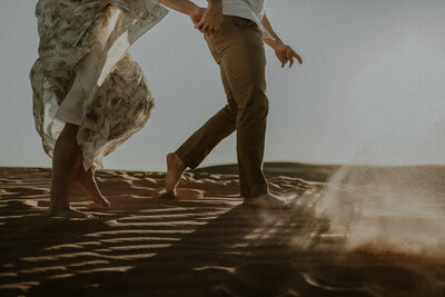Detail shot of a couples feet walking around the sand dunes while the sun is reflecting off the sand being kicked up in front of them.