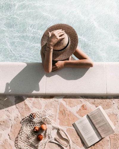 A woman relaxing by a pool holding a sun hat over her face, with a net bag, cherries, and an open book on stone tiles nearby
