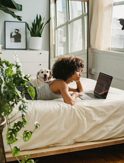 A woman and her dog laying on a bed and looking at her laptop