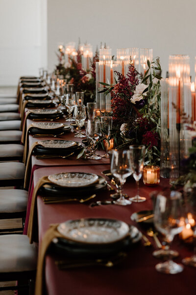 A long wedding reception table set up with moody colored decorations, flowers, and candles. 