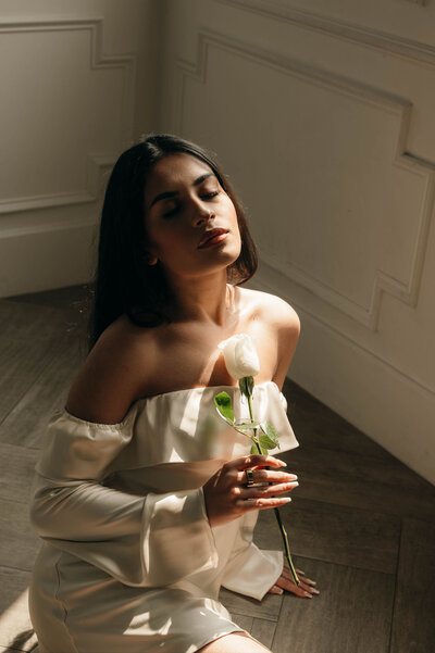 A woman in an off-shoulder white dress holds a white rose, bathed in soft sunlight. Her eyes are closed, conveying a serene and contemplative mood.