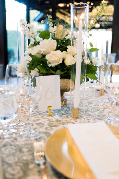 white flowers in a brass vase sitting on a table with gold plates and tall candlesticks