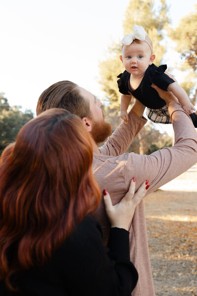 Father lifting baby girl into the air while mother looks on, captured outdoors in natural light. Photographed by Yucaipa family photographer Kaitlyn Dawn Photography.