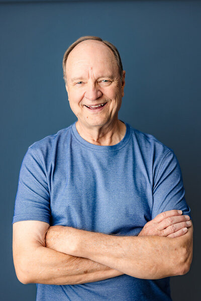 Older gentleman in blue folds arms on blue backdrop.