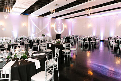 A modern reception setup at SoHo63, a Chandler wedding and event venue, featuring round tables with black linens, silver chiavari chairs, and a glowing “Mr. & Mrs.” monogram projected on the wall. Draped white fabric and soft lighting add to the upscale, romantic atmosphere.