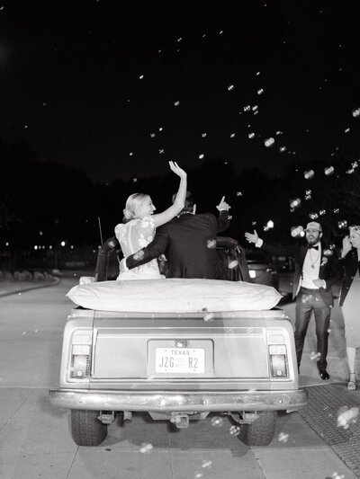 Black and white photo of bride and groom in their getaway car waving and surrounded by bubbles