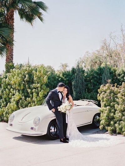 Kohnur Chic couple with bride in natural glowing makeup and a sleek low bun wearing a lace gown, posing by a vintage Porsche at an estate driveway Californian Wedding
