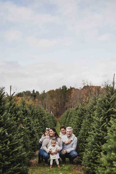 Family of five crouching together between rows of evergreen trees during a Christmas mini session at a tree farm in Northeast Ohio, with autumn foliage in the background.
