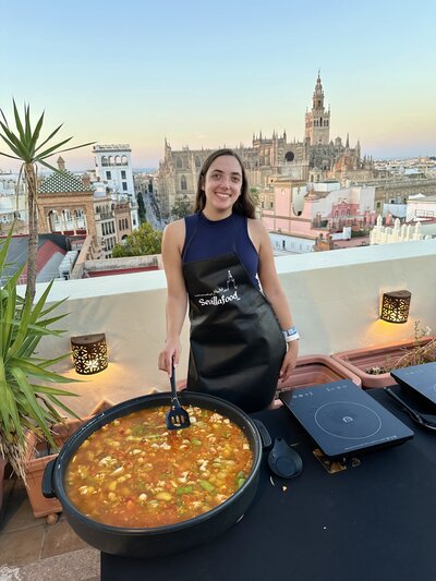 A rooftop paella cooking class in Seville, Spain - me standing cooking paella, with a gorgeous city scape of Seville behind me at sunset