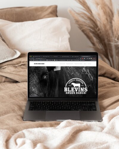 An open laptop sits on top of a bed made with tan linen bedding and displays the homepage for a Montana cattle ranch called Blevins Angus Ranch